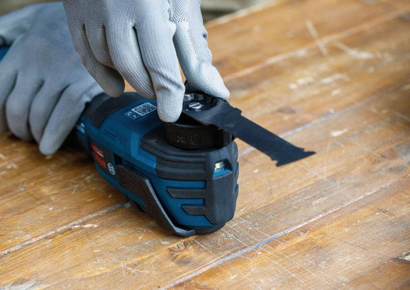 Person mit Handschuhen, der das Blatt eines elektrischen Multifunktionswerkzeugs wechselt, das auf einer Holzoberfläche platziert ist.