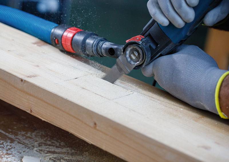 Eine Person mit Handschuhen benutzt eine elektrische Säge, um in ein Holzbrett zu schneiden und erzeugt dabei Sägespäne. Ein blauer Staubsaugerschlauch sammelt die Trümmer auf.
