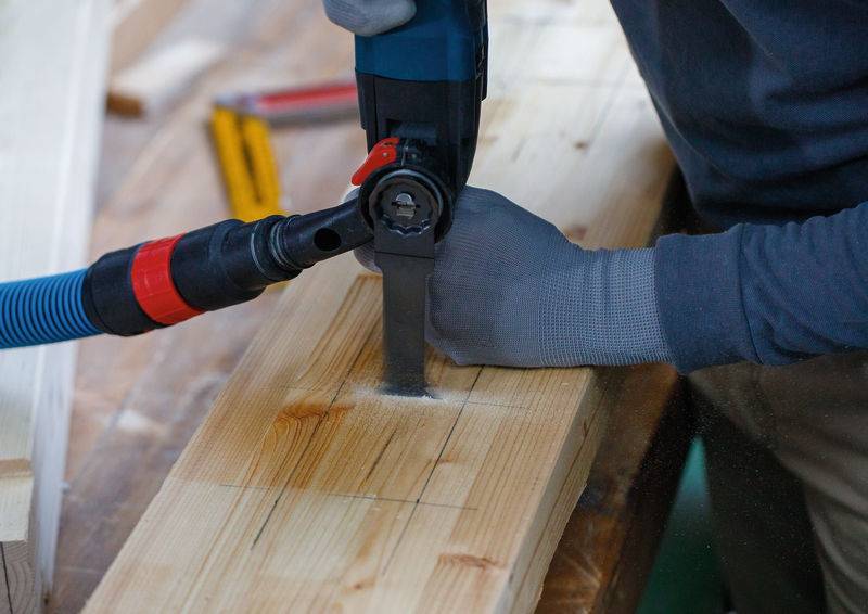 Eine Person in grauen Handschuhen nutzt ein Elektrowerkzeug mit Staubsaugeranschluss, um ein Holzbrett zu schneiden, auf einer Werkbank mit Messwerkzeugen in der Nähe.