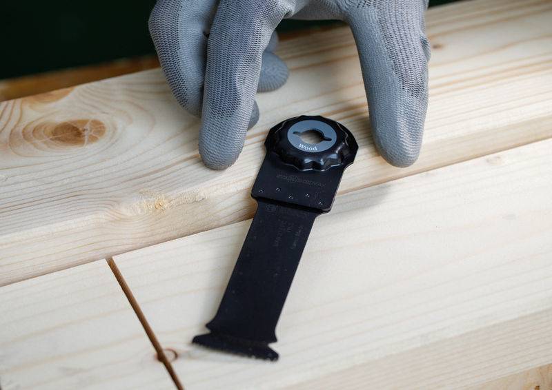Eine Hand in einem grauen Handschuh zeigt auf ein oszillierendes Multifunktionswerkzeug-Sägeblatt, das auf hellfarbigen Holzbrettern ruht.