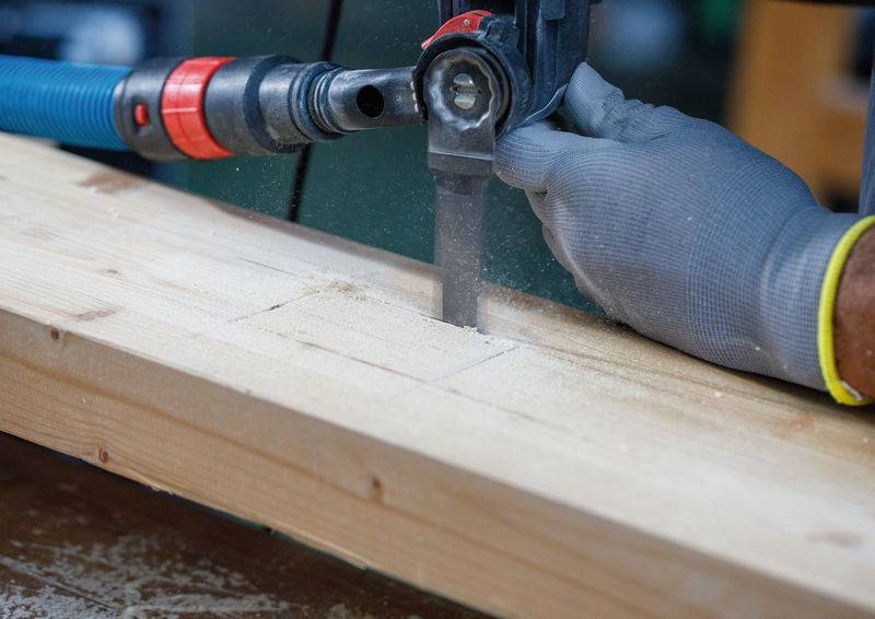 Eine behandschuhte Hand benutzt eine elektrische Säge, um ein Stück Holz zu schneiden, wobei Sägespäne sichtbar sind. Die Szene deutet auf eine Holzbearbeitungs- oder Bauaufgabe hin.