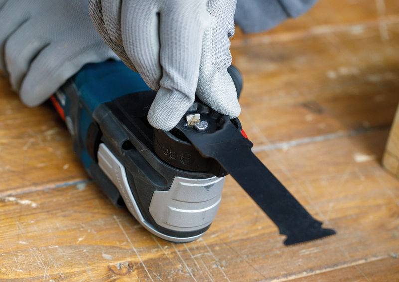 Eine Person mit grauen Handschuhen benutzt ein oszillierendes Multifunktionswerkzeug auf einer Holzoberfläche und stellt die Klinge für präzises Schneiden ein.