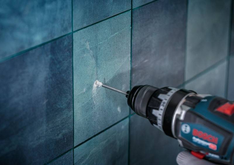 Ein Bohrer erzeugt ein Loch in einer blaukeramischen Wand und produziert sichtbaren Staub. Der Bohrer dringt in die Oberfläche ein und zeigt laufende Bohrarbeiten an.