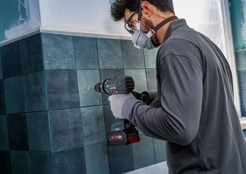 Eine Person mit Schutzbrille und Maske bohrt konzentriert in eine dunkle geflieste Wand. Sie hält einen kabellosen Elektroschrauber.