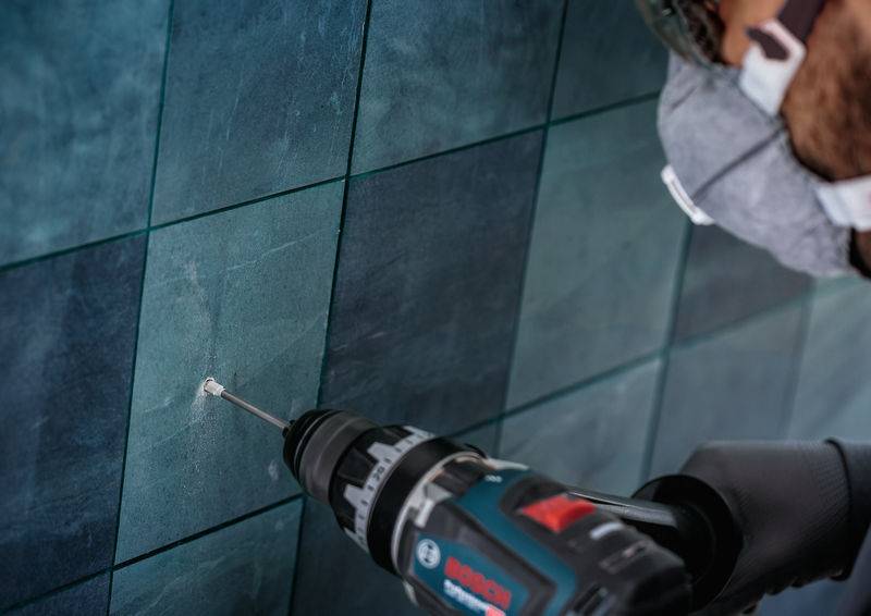 Eine Person bohrt in eine blaufliesige Wand und trägt dabei eine Schutzmaske und Schutzbrille. Der Bohrer ist schwarz und die Fliesen haben eine glänzende Oberfläche.