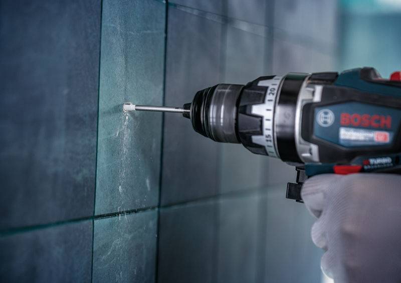 Eine Hand in einem Handschuh bedient einen Bosch-Elektroschrauber, um in eine geflieste Wand zu bohren, wobei weißer Staub um den Bohrmeißel herum auftritt.