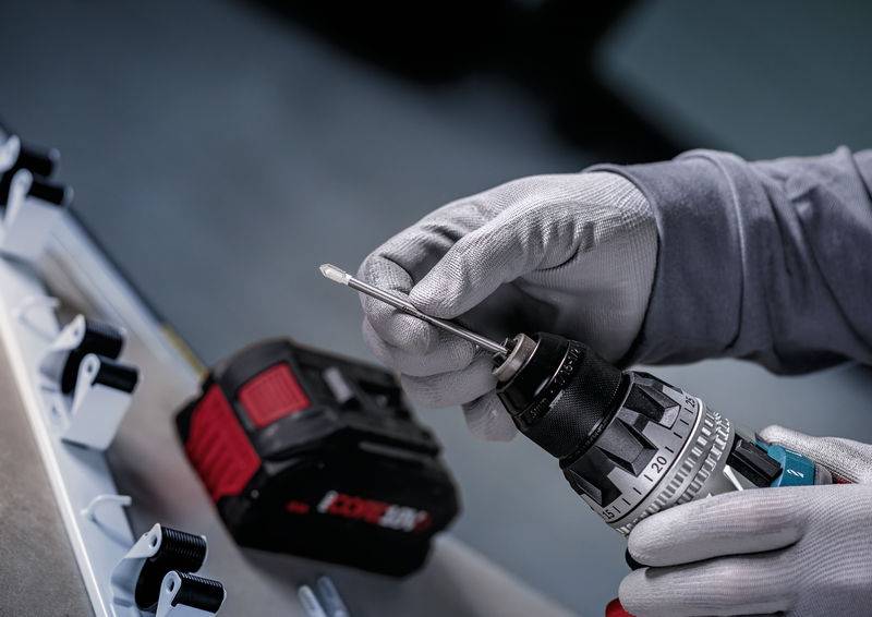 Eine Person, die Handschuhe trägt, verwendet einen Akkubohrer auf einer metallischen Oberfläche, mit einer sichtbaren roten und schwarzen Batterie neben dem Bohrer.