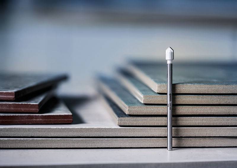 Ein Bohrbit steht vertikal neben einem Stapel von Keramikfliesen unterschiedlicher Dicken, der verschiedene Fliesenschichten hervorhebt.