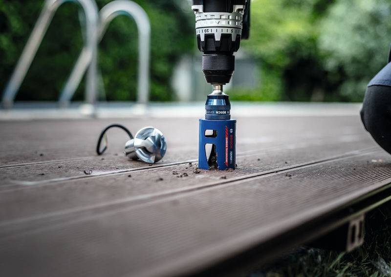 Ein Elektroschrauber mit einer blauen Lochsäge schneidet durch eine Holzterrasse, während eine andere Aufsatzvorrichtung in der Nähe liegt, vor einem unscharfen Außenhintergrund.