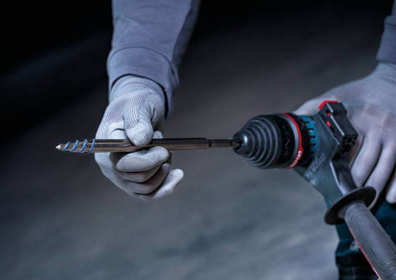Eine Person trägt Handschuhe und hält einen Bohrer mit einem Metallbohrer in der Hand, um das Werkzeug zu benutzen. Der Hintergrund ist verschwommen und dunkel.