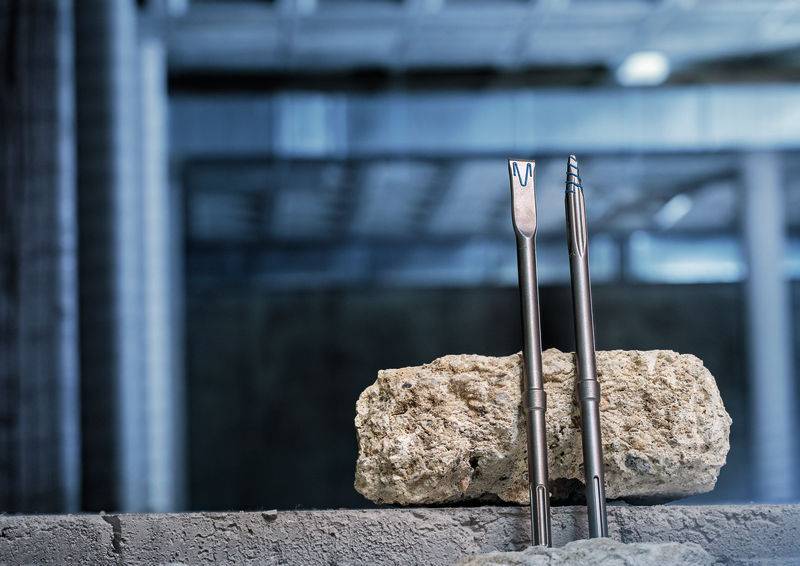 Zwei Betonmeißel stehen vertikal neben einem rauen Steinblock in Innenräumen, mit unscharfem Industriehintergrund.