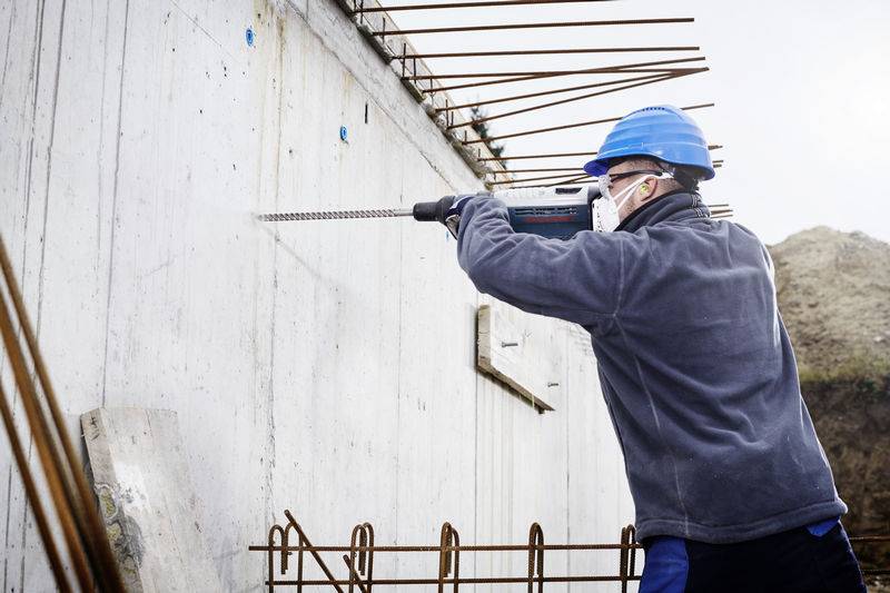 Ein Bauarbeiter in einem blauen Helm und einer Maske bedient einen Elektroschrauber an einer Betonwand auf einer Baustelle.