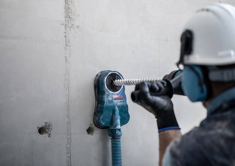 Ein Arbeiter in Schutzausrüstung benutzt eine Elektrobohrmaschine an einer Betonwand, wobei Staubabsaugungsgeräte sichtbar sind.