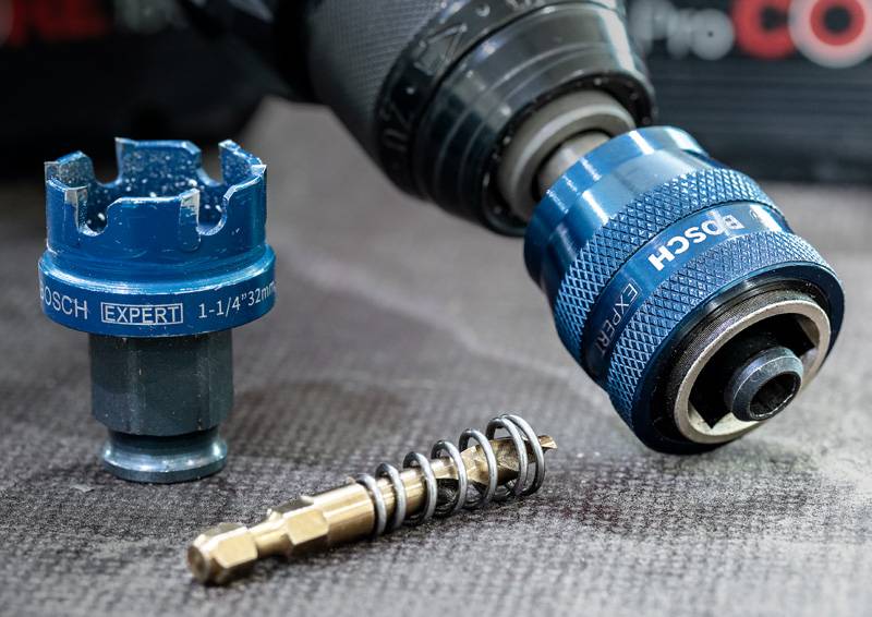 Ein Bohraufsatz und eine Lochsäge liegen auf einer grauen Oberfläche mit mechanischen Komponenten und zeigen die Werkzeuge zum Bohren und Schneiden.