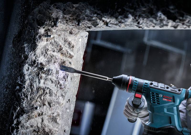 Nahaufnahme eines handgesteuerten Elektrowerkzeugs, das durch eine Betonoberfläche meißelt und in einer Industrieumgebung Staub und Trümmer erzeugt.