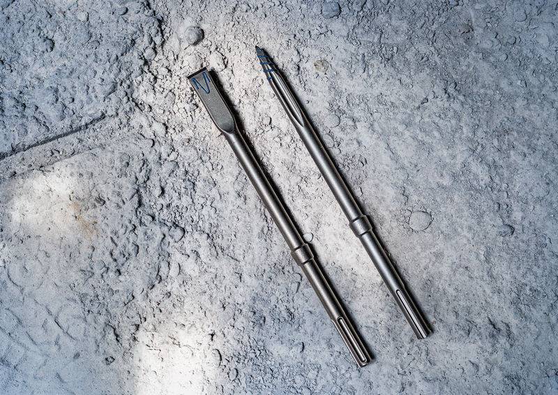 Zwei Metallbohrer auf einer staubigen Betonoberfläche, einer mit einer Meiselspitze und der andere mit einer spitzen Spitze, verwendet für schwere Aufgaben.