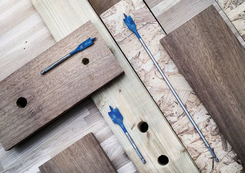Holzbretter mit gebohrten Löchern, zwei blaue Bohrer verschiedener Größen auf den Brettern platziert, auf einem hellem Holzhintergrund.