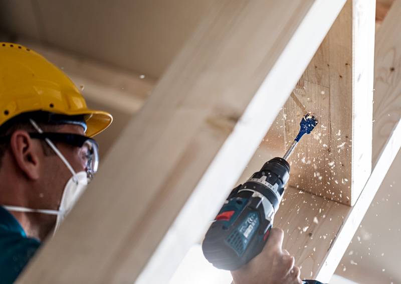 Eine Person in Schutzausrüstung verwendet eine Elektrobohrmaschine an Holzbalken und erzeugt dabei Holzspäne.
