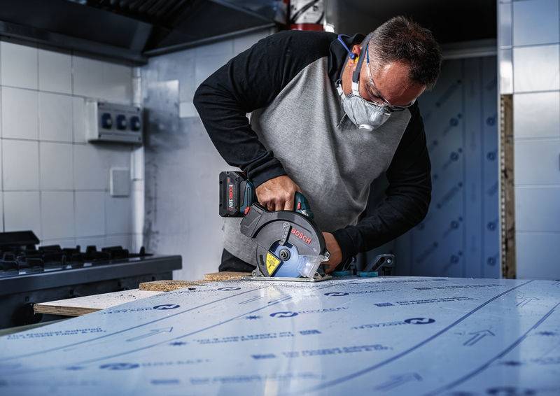 Eine Person, die Schutzbrille und Maske trägt, verwendet eine Kreissäge, um ein Materialblatt in einer Werkstattumgebung zu schneiden.