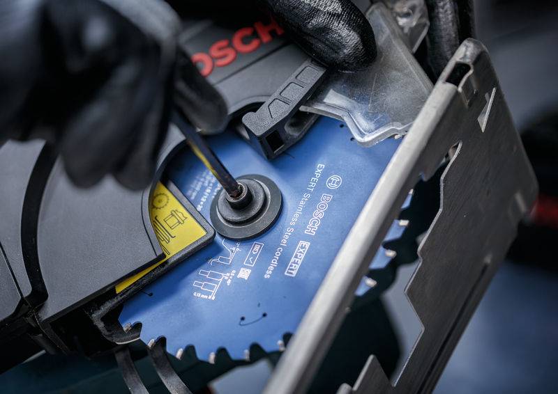 Eine Person benutzt ein Werkzeug, um ein Sägeblatt einer Bosch-Kreissäge einzustellen, wobei der Fokus auf dem Blattmechanismus und den gekennzeichneten Sicherheitsanweisungen liegt.