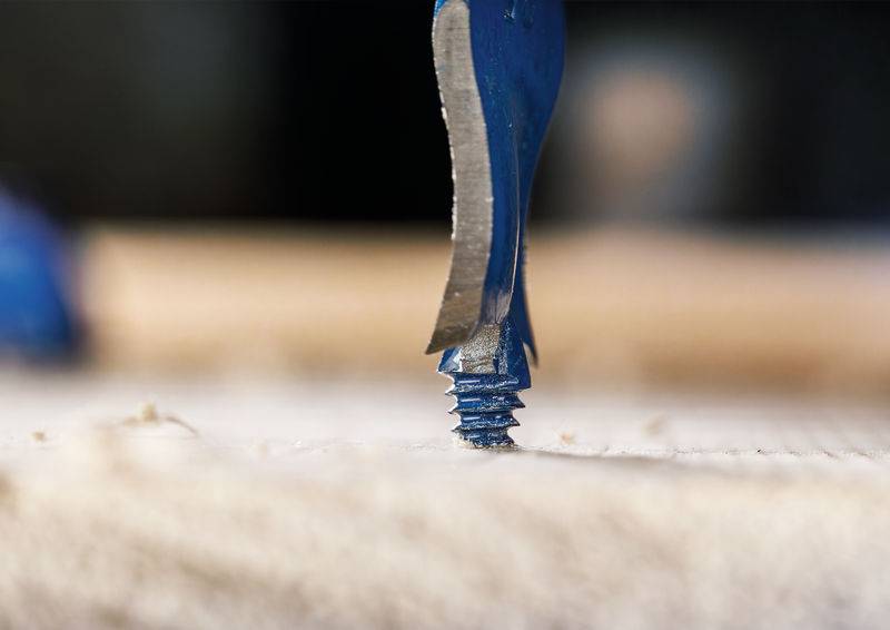 Ein blauer Bohrer dringt in eine Holzoberfläche ein und erzeugt dabei Holzspäne während des Bohrens. Nahaufnahmeansicht, die die scharfe Spiralkante des Werkzeugs zeigt.
