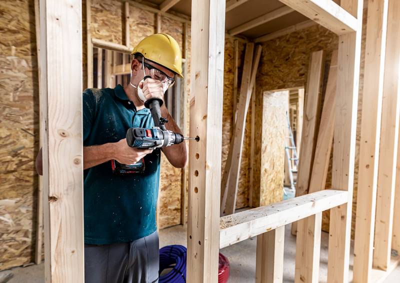 Arbeiter mit Schutzhelm, der eine Elektrobohrmaschine an einem Holzrahmen in einem Gebäude unter Konstruktion verwendet und dabei Sicherheit und Bauprozess hervorhebt.