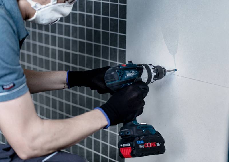 Eine Person, die eine Gesichtsmaske und Handschuhe trägt, benutzt einen Elektroschrauber, um ein Loch in die Fliesenwand zu bohren, was auf eine Bau- oder Reparaturaufgabe hindeutet.