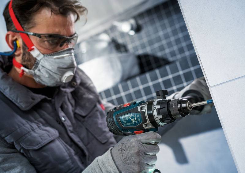 Eine Person in Schutzausrüstung verwendet einen Elektroschrauber an einer gekachelten Wand. Sie trägt Schutzbrille, Maske, Gehörschutz und Handschuhe.