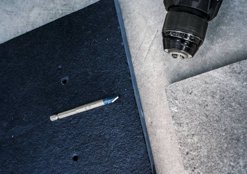 Ein Bohrer und ein Bohrbit auf einer grauen Oberfläche mit zwei Materialpièces, eines schwarz mit gebohrten Löchern und eines grau, was auf laufende Installationsarbeiten hindeutet.