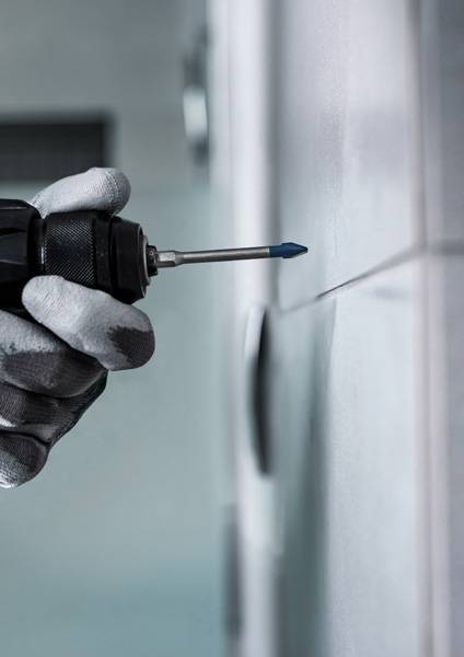 Eine Hand in einem Handschuh hält einen Bohrer gegen eine geflieste Wand und bereitet sich darauf vor, in die Fliese zu bohren.