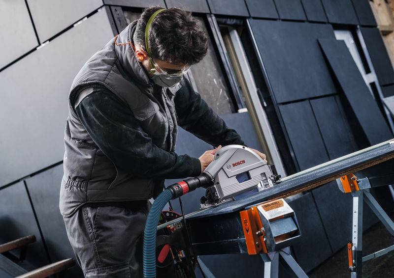 Eine Person in Schutzausrüstung benutzt eine Kreissäge, um ein großes Brett zu schneiden, und demonstriert dabei Holzbearbeitungs- oder Bauarbeiten.