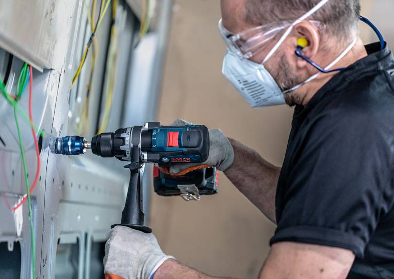 Eine Person in Schutzausrüstung benutzt einen Elektroschrauber an einer Wand mit sichtbarer Verdrahtung, was auf Bau- oder Elektroinstallationsarbeiten hindeutet.