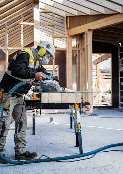 Ein Bauarbeiter in Schutzausrüstung benutzt eine Kreissäge an einem Holzbalken auf einer Baustelle mit Holzrahmenkonstruktionen im Hintergrund.