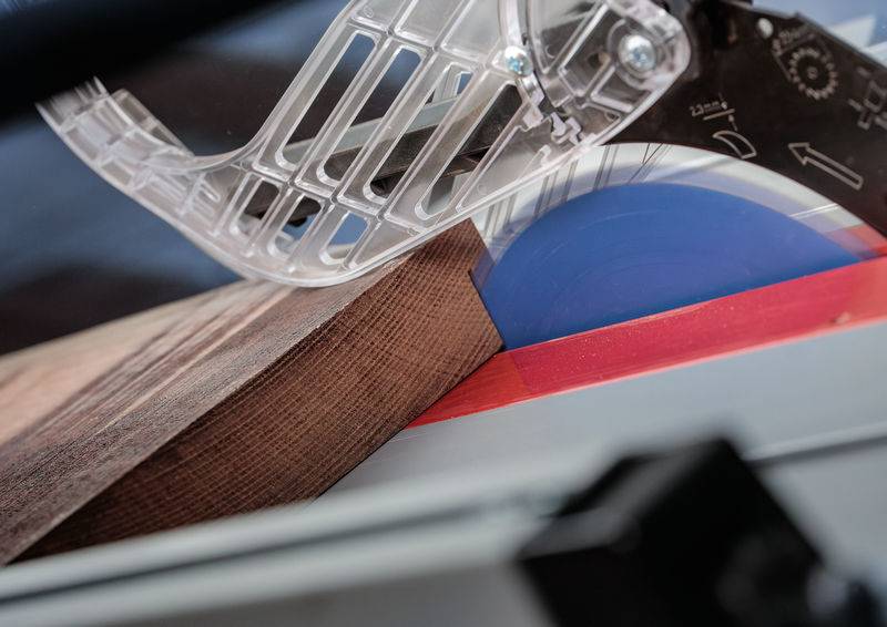 Eine Nahaufnahme einer Tischkreissäge, die ein Stück Holz in einem Winkel schneidet, mit einer transparenten Schutzvorrichtung, die das blaue Sägeblatt teilweise abdeckt.