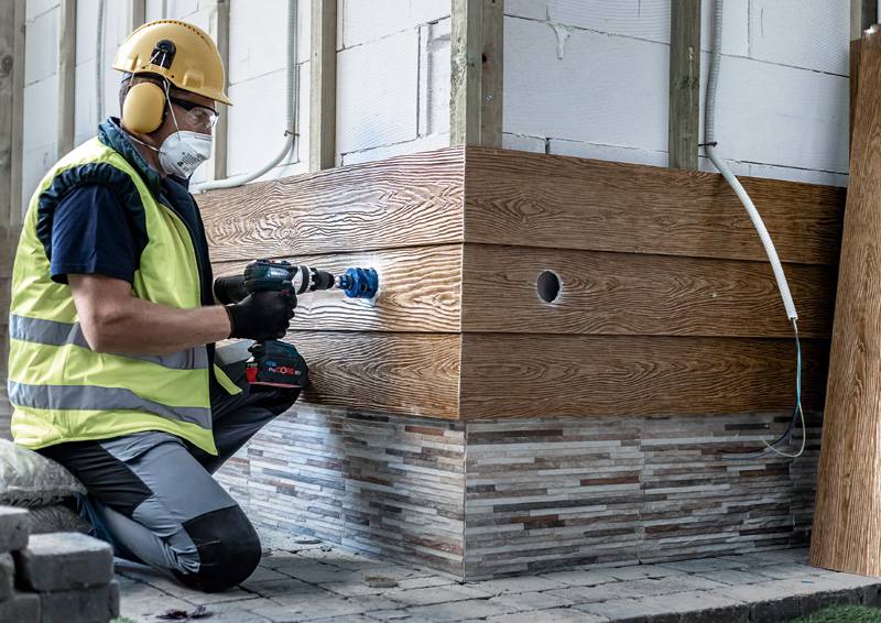 Arbeiter in Schutzausrüstung bohrt in die Holzverkleidung eines Gebäudes, was auf Bau- oder Renovierungstätigkeit hindeutet.