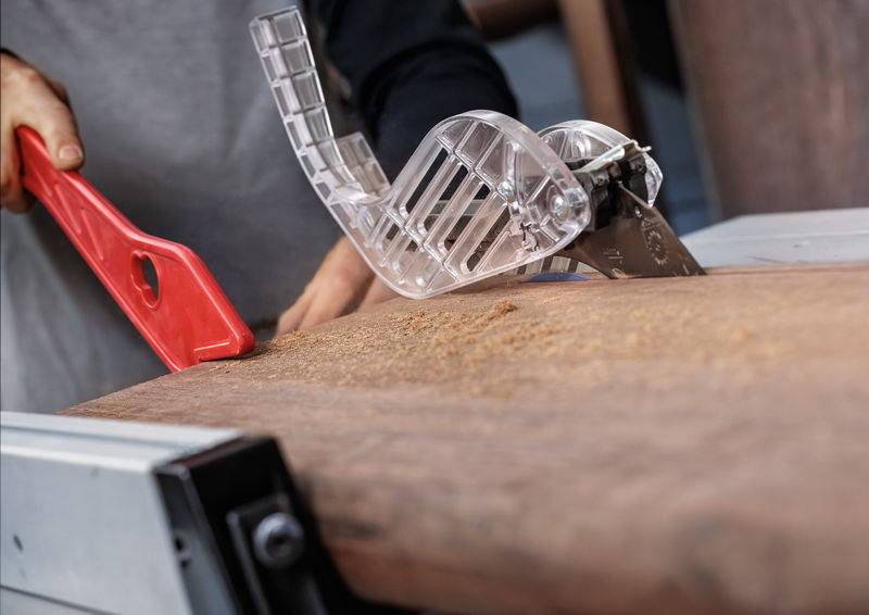 Eine Person bedient eine Tischkreissäge mit transparenter Schutzvorrichtung und schneidet ein Holzbrett. Sicherheitsausrüstung ist sichtbar und unterstreicht den sorgfältigen Umgang.