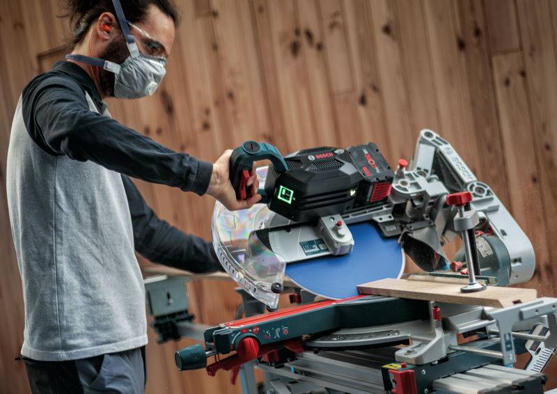 Eine Person benutzt eine Kappsäge, um ein Stück Holz in einer Werkstatt zu schneiden, und trägt dabei eine Schutzbrille und eine Staubmaske zum Schutz.