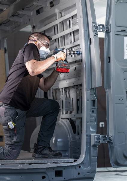 Ein Arbeiter in Schutzausrüstung verwendet einen Bohrer im Inneren eines Transporters und konzentriert sich auf die Installation oder Reparatur von Hardwarekomponenten.