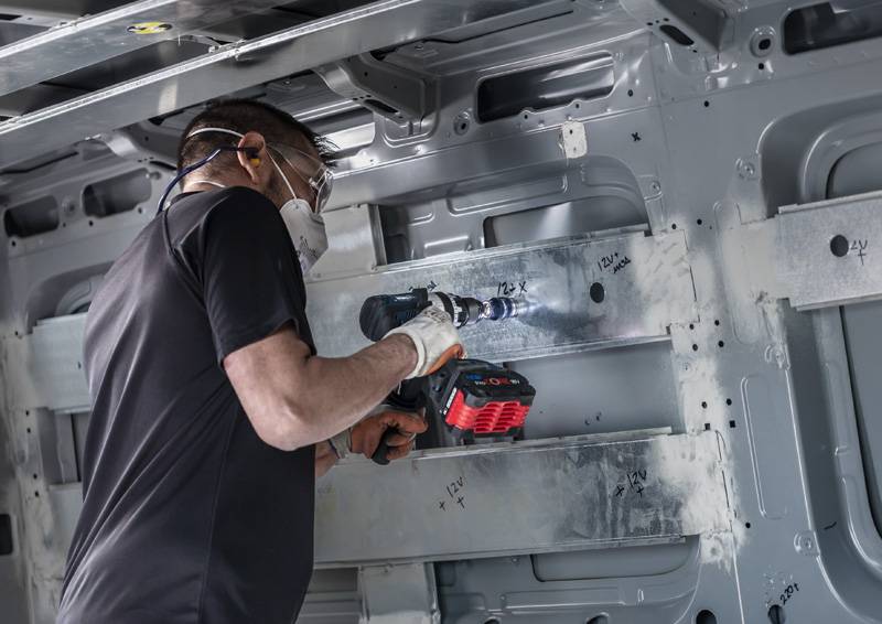Eine Person in Schutzausrüstung verwendet einen Elektroschrauber, um an der Metallinnenseite eines Fahrzeugs zu arbeiten und konzentriert sich dabei auf die Befestigung von Metallhaltern.