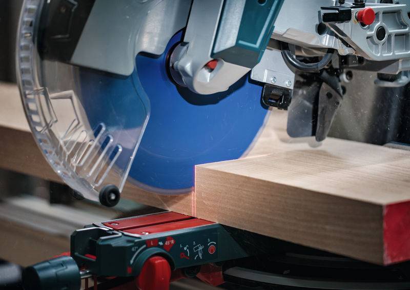 Eine Nahaufnahme einer Gehrungssäge, die durch ein Holzbrett schneidet, wobei die blaue Sägeblatt und die Laserführung für präzises Schneiden hervorgehoben werden.