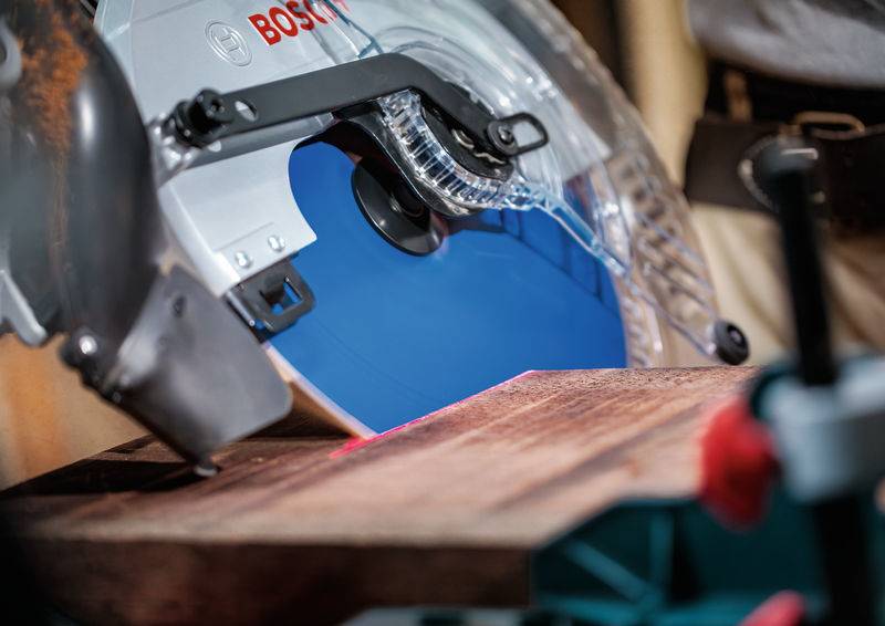 Eine Kreissäge mit einem blauen Blatt schneidet ein Stück Holz auf einer Werkbank, das an Ort und Stelle eingespannt ist. Eine Schutzvorrichtung ist sichtbar.