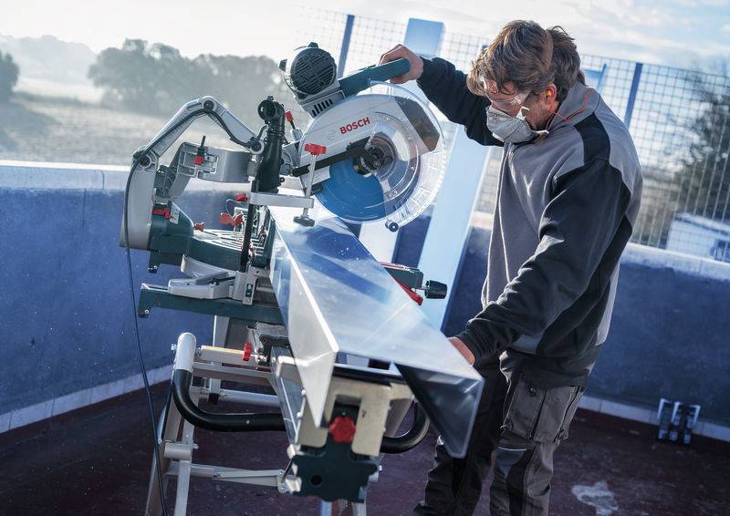 Ein Mann, der eine Schutzmaske trägt, nutzt eine Gehrungssäge, um einen Metallbalken an einem klaren Tag im Freien zu schneiden.