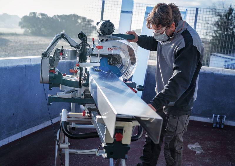 Eine Person bedient eine Gehrungssäge, um einen langen Metallbalken im Freien zu schneiden. Sie tragen eine Schutzbrille und eine Maske zum Schutz, mit Morgenlicht im Hintergrund.