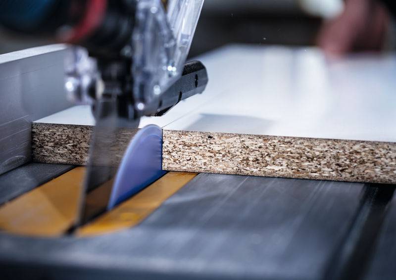 Eine Nahaufnahme einer Tischkreissäge, die durch ein Holzbrett schneidet, zeigt das Sägeblatt in Aktion und Sägespanpartikel in der Luft.