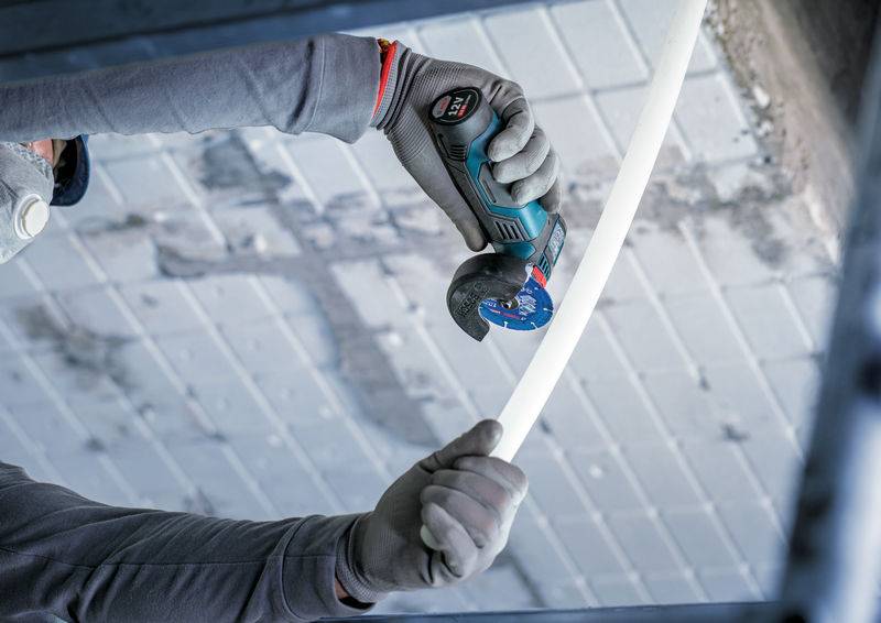 Eine Person benutzt ein tragbares Rotationswerkzeug, um ein weißes Kunststoffrohr gegen eine graue geflieste Oberfläche zu schneiden, was auf eine Bau- oder Reparaturaktivität hindeutet.