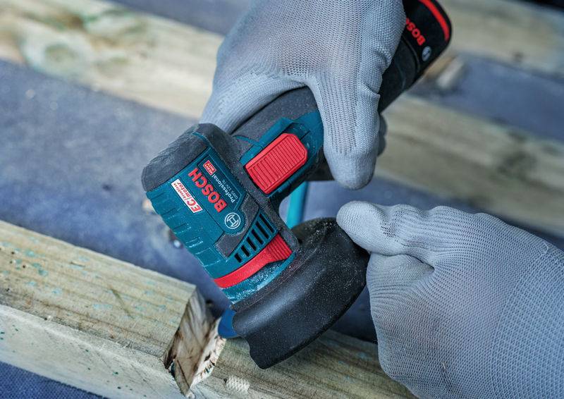 Eine Person mit Handschuhen benutzt einen Bosch-Elektroschleiffer auf einer Holzoberfläche und hebt dabei das blaue und rote Design des Werkzeugs sowie den ergonomischen Griff hervor.