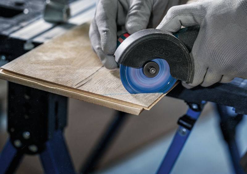 Hände mit Handschuhen verwenden ein Elektrowerkzeug, um ein Stück Holz zu schneiden.