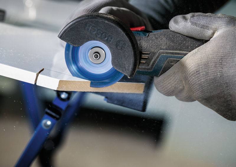 Eine Person trägt Handschuhe und benutzt ein Elektrowerkzeug mit einer kreisförmigen Klinge, um ein Stück Holz zu schneiden, wobei dabei Sägespäne entstehen.