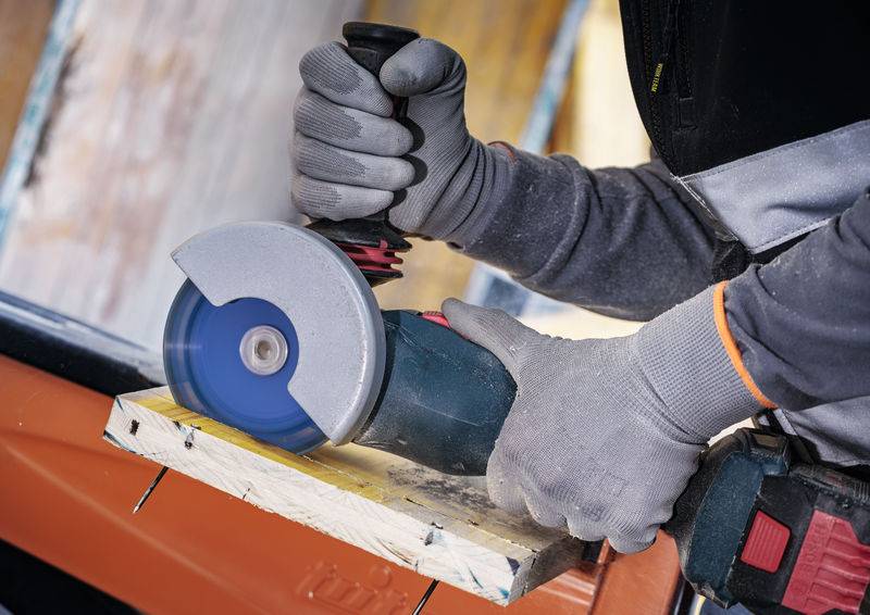 Eine Person, die eine blaue Kreissäge verwendet, um ein Stück Holz zu schneiden, während sie zum Schutz Schutzhandschuhe und Schutzkleidung trägt.
