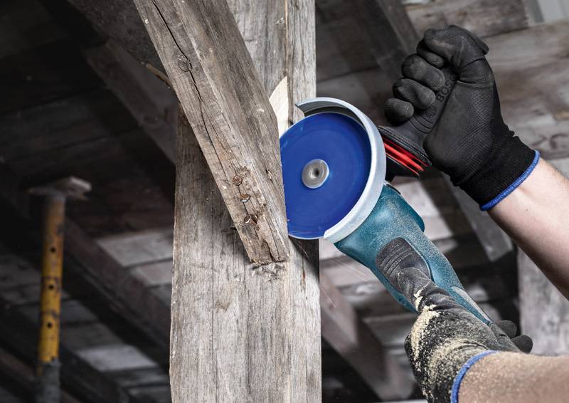 Eine Person mit Handschuhen verwendet einen blau-silbernen Winkelschleiffer, um einen Holzbalken in einer Werkstatt zu schneiden.
